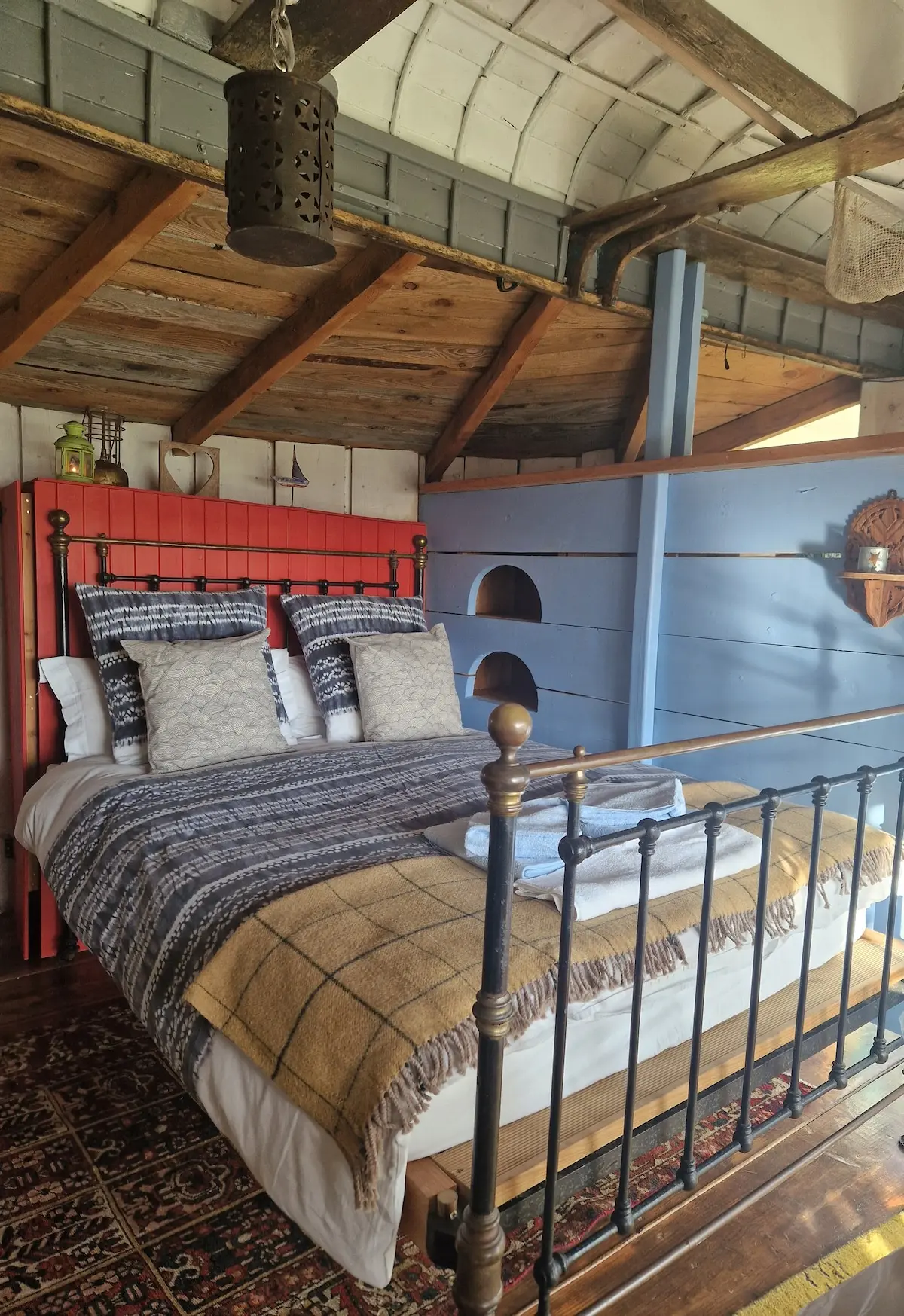 The iron bed dressed with throws and cushions, red headboard, blue boat-hull wall behind, and lantern above