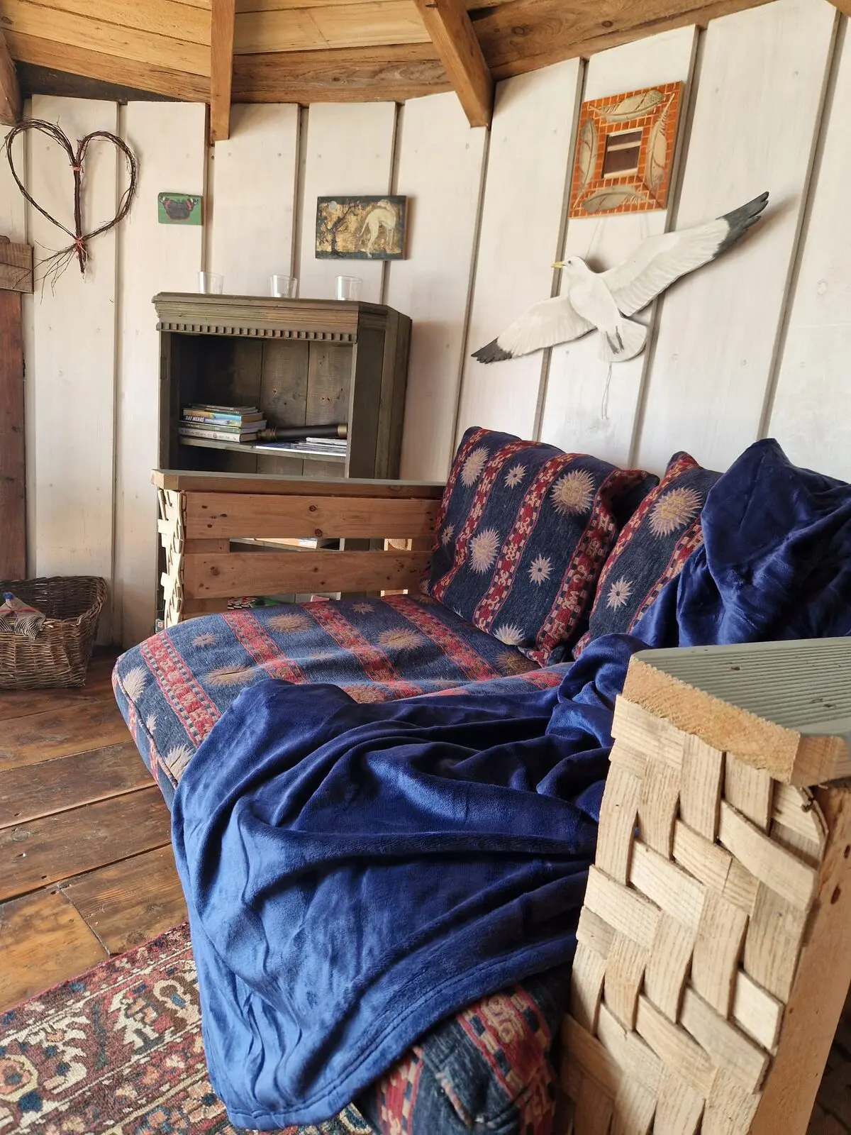 The sofa corner with patterned cushions and blue throw, kittiwake sculpture on the wall, bookshelf and heart decoration